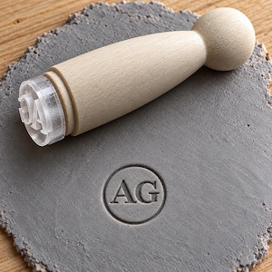 Puede incluir: Un sello de madera clara con mango redondeado y base de acrílico transparente que muestra las letras AG. El sello se sitúa sobre un disco circular de arcilla gris que presenta una impresión en relieve de las letras AG dentro de un círculo.