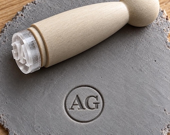 May include: A light-colored wooden stamp with a rounded handle and a clear acrylic base featuring the letters AG. The stamp is positioned above a circular gray clay disc that displays a debossed impression of the letters AG inside a circle.