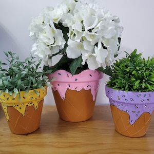 Könnte beinhalten: Drei bemalte Terrakotta-Blumentöpfe in Form von Eiswaffeln. Die Töpfe sind mit gelbem, rosa und lila Eis und Streuseln verziert. Die Töpfe sind mit grünen und weißen Pflanzen gefüllt.