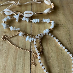 May include: Three layered necklaces with white shell and bead accents. The top necklace is made of white shells and string, the middle necklace is made of white beads and gold chain, and the bottom necklace is made of white beads and gold chain.