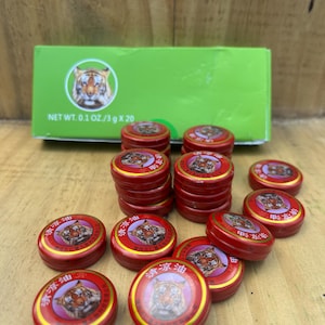 以下が含まれることがあります： タイガーバームの赤い丸い金属製の容器が20個入った箱。各容器には、虎のイラストと中国語の文字が書かれた赤と金のラベルが付いています。箱は緑色で、虎のイラストと「NET WT. 0.1 OZ/3 g X 20」の文字が書かれています。
