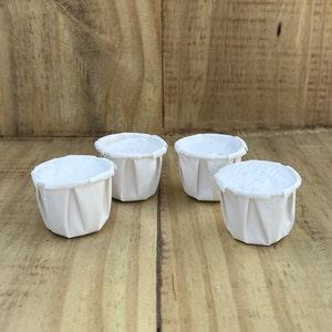 May include: Four white paper cups, each with a fluted design, are arranged in a row on a wooden surface.