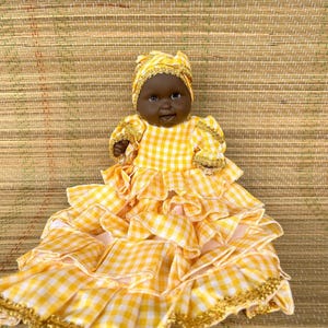 May include: A handcrafted baby doll with dark skin, wearing a yellow and white gingham dress with ruffled layers and gold trim. The doll also has a matching head wrap with gold accents. The doll is a charming example of folk art.