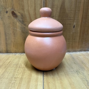 Puede incluir: Un pequeño tarro de terracota con tapa. El tarro es de color naranja claro y tiene una forma redonda y lisa.