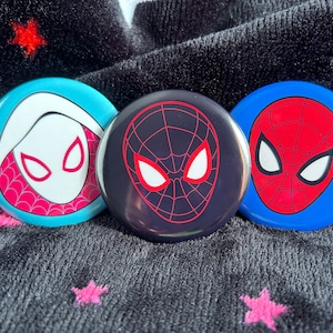 May include: Three round pinback buttons featuring different Spider-Man characters. The first button is blue with a white and pink Spider-Gwen design. The second button is black with a red Spider-Man design. The third button is blue with a red Spider-Man design.