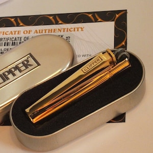 May include: A gold-colored Clipper lighter and poker set in a silver metal case. The lighter and poker are displayed on a black velvet-like surface. The case is open, revealing the contents. The word "CLIPPER" is printed on the lighter and case.