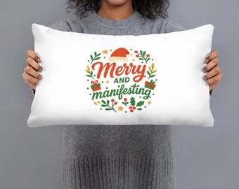 Puede incluir: Cojín rectangular blanco con el texto "Merry and Manifesting" en rojo y verde, rodeado de ilustraciones festivas que incluyen un gorro de Papá Noel, regalos y acebo. El cojín lo sostiene una persona.