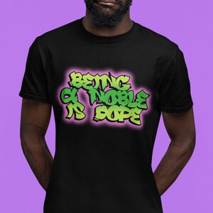 以下が含まれることがあります： 「Being a Noble is Dope」と書いてある緑とピンクのグラフィックがプリントされた黒いTシャツです。