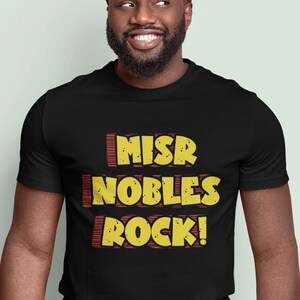 以下が含まれることがあります： 「MISR NOBLES ROCK!」という黄色と赤のグラフィックがプリントされた黒いTシャツ