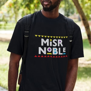 以下が含まれることがあります： カラフルなブロック体で「MISR NOBLE」と書かれた黒いTシャツ。テキストは黄色、赤、青の三角形の縁取りで囲まれています。