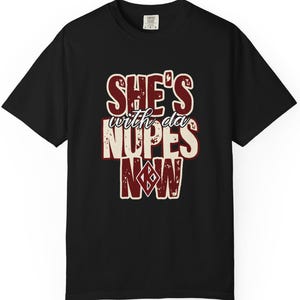 Może przedstawiać: Czarny t-shirt z napisem "SHE'S with da NUPES NOW" w postarzałej, bordowej i kremowej czcionce. Projekt zawiera emblemat w kształcie rombu. Koszulka ma okrągły dekolt i krótkie rękawy.