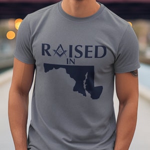 May include: A gray t-shirt with the text "RAISED IN" and a graphic of the state of Maryland with a Masonic symbol above the text.