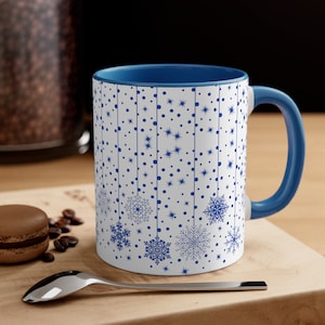 May include: A white ceramic mug with a blue rim and handle. The mug features a repeating pattern of snowflakes, stars, and dots in blue on a white background.