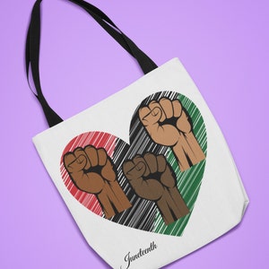 May include: A white tote bag with black handles features a heart-shaped design with three raised fists in the colours of the Pan-African flag. The text "Juneteenth" is printed below the heart.