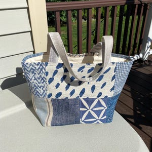 May include: A tote bag constructed from various blue and white fabric patches featuring diverse patterns including geometric shapes, brush strokes, and woven textures. The bag has two light gray fabric handles and a cream-colored interior lining.