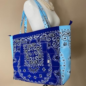 May include: A large tote bag with a blue bandana print. The bag features a patchwork design with a dark blue center and light blue accents. The straps are light blue with a white paisley pattern.