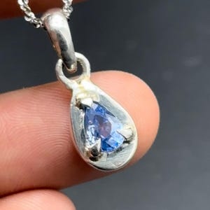 Op de afbeelding: Een zilveren hanger met een peervormige blauwe edelsteen in een gladde metalen zetting, hangend aan een fijne zilveren ketting. De hanger heeft een druppelvormig frame dat de gefacetteerde blauwe steen vasthoudt met drie kleine metalen pootjes.