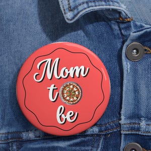 Puede incluir: Un botón rojo con el texto "Mom to be" en letra cursiva blanca y una pizza con mozzarella y albahaca encima.