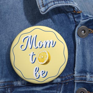 May include: A yellow button with a blue border and the text "Mom to be" in blue script. A yellow lemon slice is centered below the text.