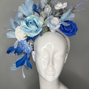 Tocado llamativo en azul hielo y zafiro real con detalles plateados y plumas, estilo Kentucky Derby.