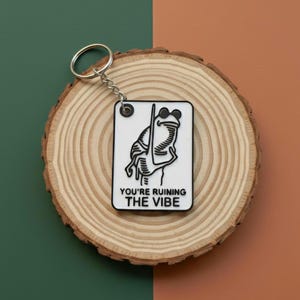 May include: A white and black keychain featuring a frog illustration holding a pole, with the text "YOU'RE RUINING THE VIBE" below. The keychain is attached to a silver metal ring and chain. The background is a wooden slice and a green and brown surface.