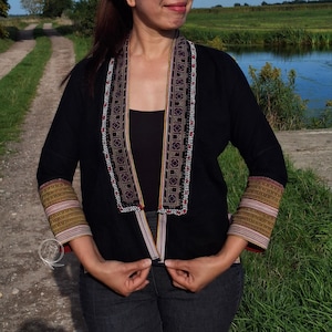 Puede incluir: Una chaqueta negra abierta con un cuello ancho que presenta un intrincado bordado geométrico en negro, blanco y rojo. Las mangas largas tienen puños anchos decorados con bandas horizontales de patrones en tonos dorados, rosas y blancos.