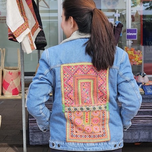 Peut inclure: Une veste en jean bleu clair avec un grand panneau rectangulaire de tissu brodé aux couleurs vives au dos. La broderie présente des motifs géométriques complexes et des motifs floraux dans des tons de rose, d'orange et de vert.
