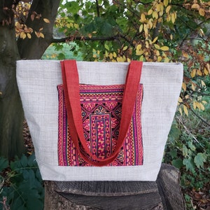 Może przedstawiać: Duża, kremowa torba typu tote z czerwonymi paskami i kolorową naszywką haftowaną z przodu. Naszywka przedstawia geometryczny wzór w odcieniach różowego, fioletowego, żółtego i czarnego.