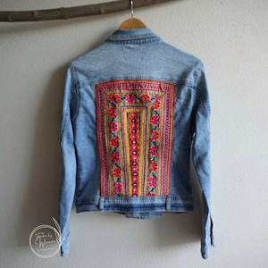 Op de afbeelding: Lichtblauwe spijkerjas met kleurrijk geborduurd bloemmotief op de rug. De borduursels bevatten roze, rode, gele en oranje bloemen. De jas hangt aan een houten hanger tegen een witte muur.