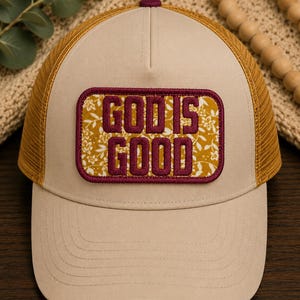 May include: A beige trucker hat with a mustard yellow mesh back and a rectangular patch on the front. The patch features a mustard yellow floral background with the maroon embroidered text "GOD IS GOOD" and a thick maroon border.