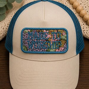 May include: Tan trucker hat with a blue mesh back and brim. The hat features a rectangular patch with the words "MADE TO WORSHIP" embroidered in blue over a floral design.