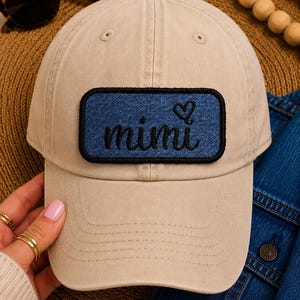 May include: A beige baseball cap with a denim patch displaying the word "mimi" in cursive and a heart symbol. The cap has a curved peak and a button on top. Sunglasses and a denim jacket are visible in the background.
