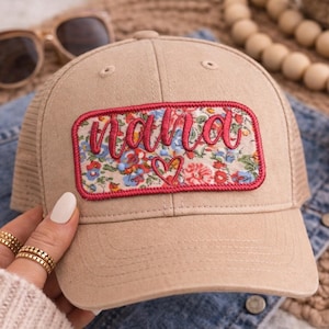 Könnte beinhalten: Beige Baseball Cap mit einem floralen "Nana" Aufnäher. Der Aufnäher hat einen roten Rand und zeigt ein Herz. Die Kappe hat einen Mesh-Rücken und einen gebogenen Schirm. Sonnenbrille und rotes Bandana im Hintergrund.