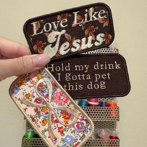 Może przedstawiać: Haftowane naszywki z kwiatowymi wzorami i tekstem. Jedna naszywka głosi "Love Like Jesus" na brązowym tle. Inna mówi "Hold my drink I gotta pet this dog". Trzecia naszywka przedstawia kokardę.