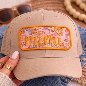May include: A beige baseball cap featuring a floral patch with the word "mimi" embroidered in yellow script. The patch has a pink background and an orange border. The cap has a mesh back and a curved peak. Sunglasses and a red bandana are in the background.