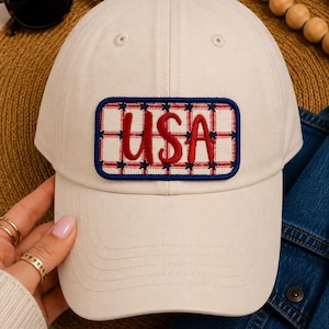 Op de afbeelding: Crèmekleurige baseballpet met een rechthoekige patch met het woord "USA" in rood schrift. De patch heeft een rood-blauw ruitpatroon met blauwe sterren. Zonnebril en een spijkerjasje zijn op de achtergrond.
