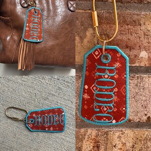 May include: A rectangular fabric keychain with rounded corners, featuring a rust-colored background with a white geometric pattern. The word RODEO is embroidered in turquoise thread, matching the turquoise border. It includes a gold-toned metal carabiner.