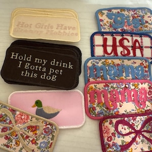 May include: A collection of embroidered patches in various shapes and colors. Some patches feature floral patterns, while others display text like "USA," "Mama," and "Hold my drink I gotta pet this dog." One patch has a duck illustration.