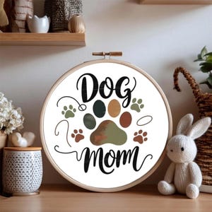 Peut inclure: Un cercle à broder avec l'inscription "Dog Mom" en écriture noire. Le motif présente une grande empreinte de patte dans des tons de vert, marron et beige, entourée de petites empreintes. Le cercle est en bois.