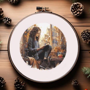 May include: A cross-stitch embroidery hoop featuring a watercolor painting of a person reading a book by a window. The scene is framed by pine needles, pine cones, and berries, creating a cozy, autumnal aesthetic.