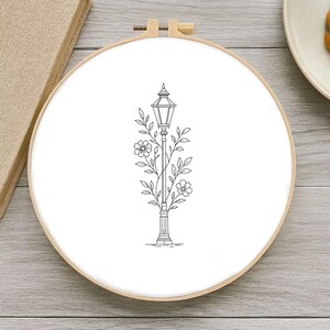 Peut inclure: Un cercle à broder avec un dessin au trait noir et blanc d'un lampadaire orné de fleurs et de feuilles. Le motif est posé sur un fond blanc à l'intérieur d'un cercle en bois, présentant un thème botanique.