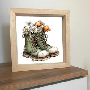 Peut inclure: Une impression encadrée présentant une illustration à l'aquarelle de bottes vertes remplies de marguerites et de fleurs orange. Les bottes ont des motifs floraux et sont lacées. L'œuvre est sur fond blanc et encadrée en bois clair.