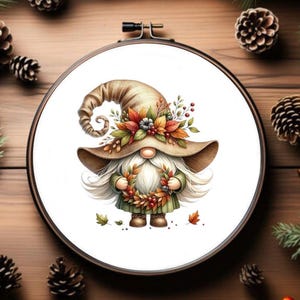 May include: An embroidery hoop featuring a gnome illustration. The gnome is depicted with a brown hat decorated with autumn leaves and berries. It has a long white beard and holds a wreath of fall foliage. The background is a wooden surface with pinecones and greenery.