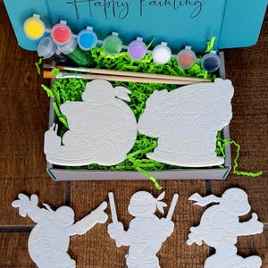 May include: A craft kit for painting featuring Teenage Mutant Ninja Turtles. The kit includes paint pots in various colors, paintbrushes, and several unpainted turtle figures. The box is teal and has the words "Happy Painting" written on it.