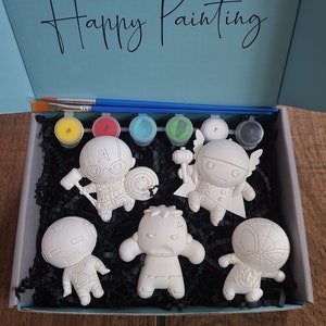 Puede incluir: Un conjunto de seis figuras de cerámica blanca de superhéroes en una caja azul con las palabras "Happy Painting" en la tapa. Las figuras están listas para ser pintadas.