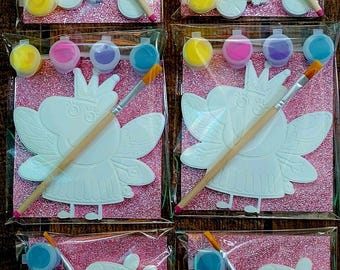 Peut inclure: Kits de peinture par numéros avec un personnage de dessin animé blanc avec une couronne et des ailes, une voiture et des pinceaux. Chaque kit comprend un fond rose pailleté et de petits pots de peinture jaunes, roses, bleus et violets.