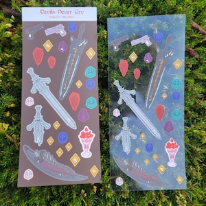 May include: Two sticker sheets featuring various designs, including swords, guns, and dessert items. The text "Devils Never Cry" is visible at the top of one sheet. The stickers are in shades of gray, brown, red, and purple.