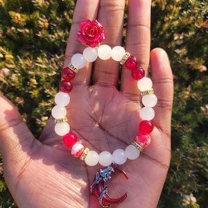 May include: A beaded bracelet featuring white and red beads, accented with gold-colored spacers. A red crescent moon charm with a dangling red gem hangs from the bracelet. A red rose adorns the top of the bracelet.