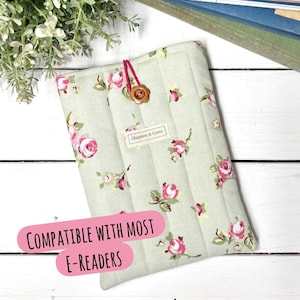 May include: A pale green quilted e-reader cover featuring a floral design of pink roses and green foliage. It has a button fastening and a label reading "Chapters & Coves". The text "Compatible with most e-readers" is also visible.