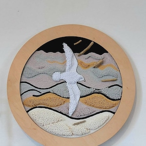 Puede incluir: Una pieza de arte de pared circular enmarcada en madera con un pájaro blanco volando sobre un paisaje texturizado de colores beige, marrón y gris.